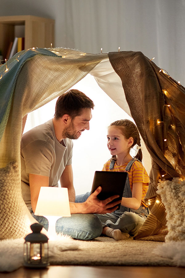 Father and daughter sitting in a blanket fort with fairy lights looking at a tablet together